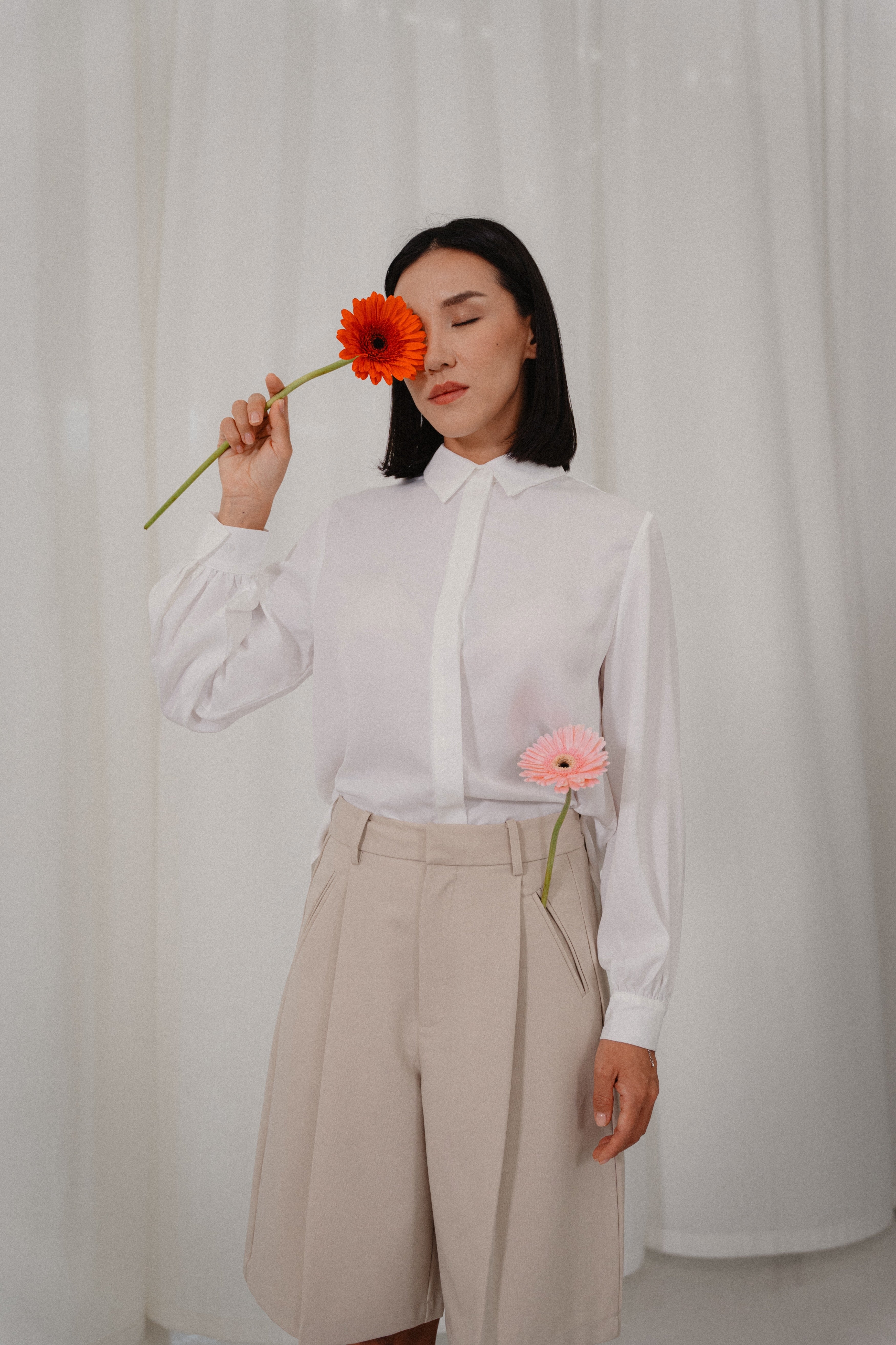 A woman wearing beige bermuda pants and white collared long-tailed blouse with puffy sleeves and a back pleated tail that creates a flowing silhouette, designed for office and city wear.