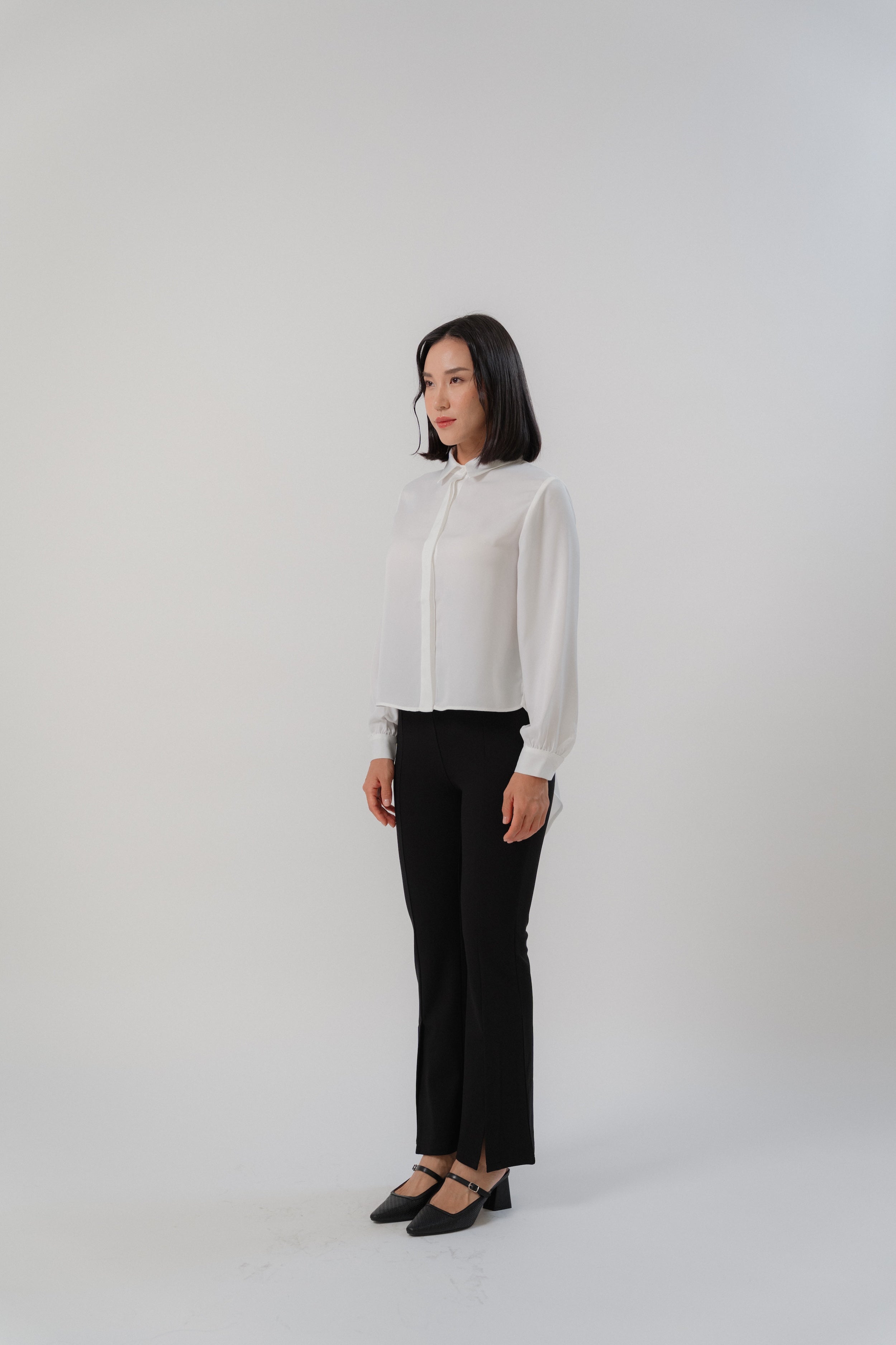 A woman wearing black bootcut pants and white collared long-tailed blouse with puffy sleeves and a back pleated tail that creates a flowing silhouette, designed for office and city wear.